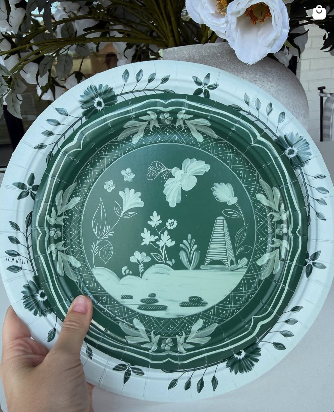 Decorative green plate with floral and landscape design held by a hand against a blurred background.